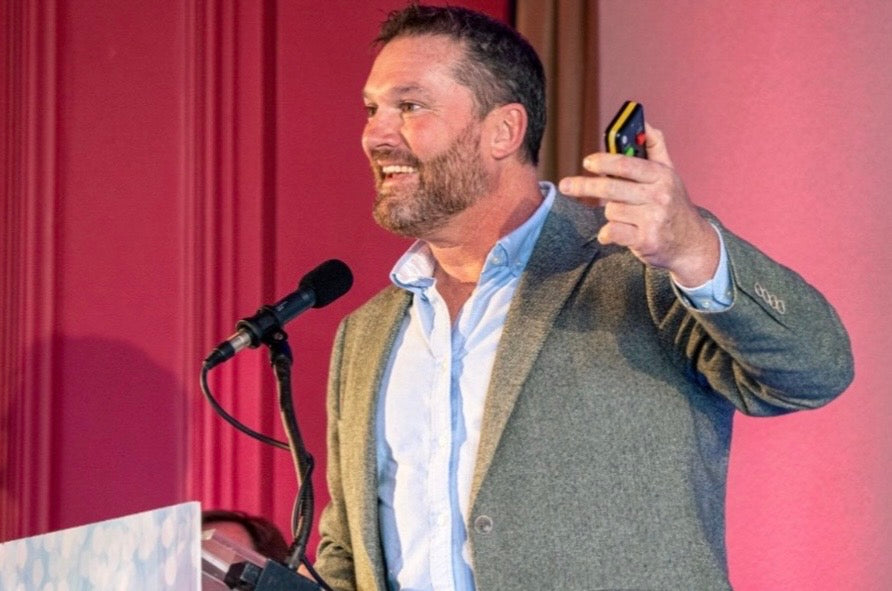 Brendon Prince speaking at a microphone and holding a presentation remote