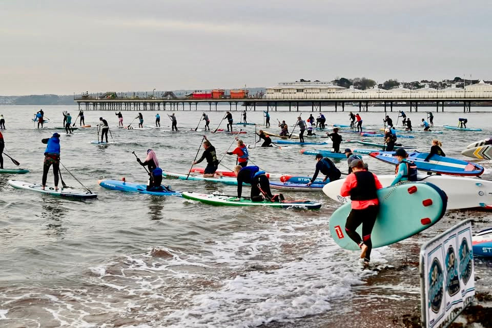 Paddleboarders entering the sea beside a pier for a coastal SUP event