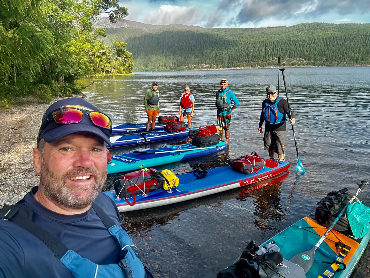 Brendon Prince and a group with paddleboards and expedition bags beside a wooded lake